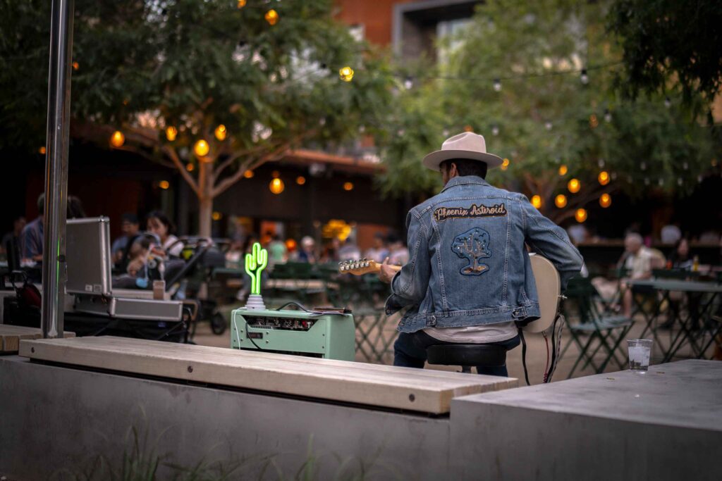 A man in a denim jacket plays guitar aside a retro amplifier with a neon cactus. Diners in the background enjoy the ambience.